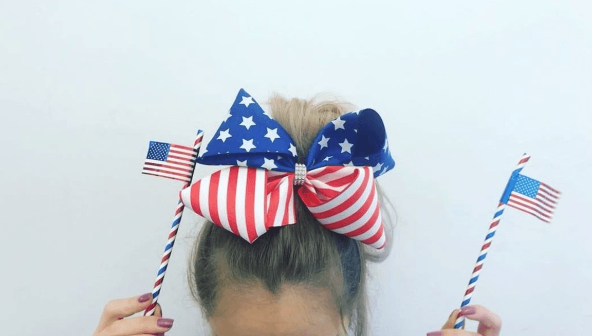 Girl wearing LuvHer Shop American flag hair bow, holding mini USA flags, patriotic accessory